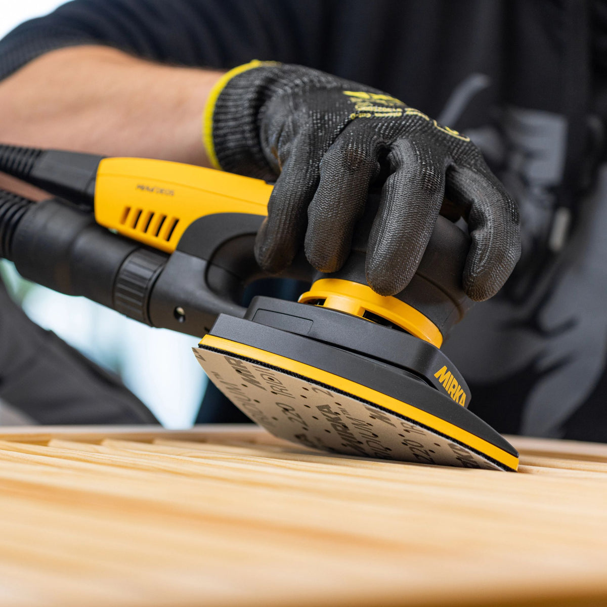 A close-up of a person wearing a black knit work glove and black shirt, using a yellow and black Mirka DEOS II Delta sander on a light wood surface.