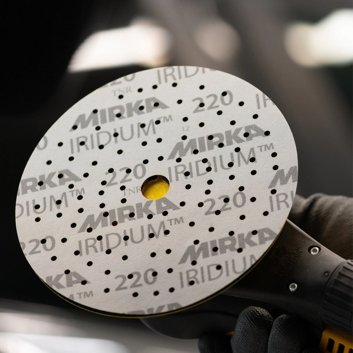 A close-up of a circular Mirka Iridium 220 sanding disc with a yellow center hole, held by a gloved hand against a blurred dark background.