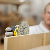 A close-up action shot of a person using a yellow and black orbital sander with a gray sanding disc on a light wood board in a workshop setting.