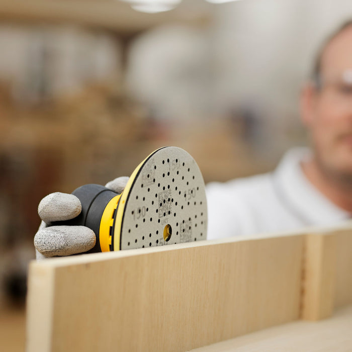 A close-up action shot of a person using a yellow and black orbital sander with a gray sanding disc on a light wood board in a workshop setting.