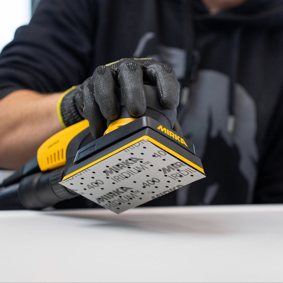 A person wearing a black hoodie and a black work glove holds a yellow and black Mirka DEOS sander tool with Iridium pad visible underneath.